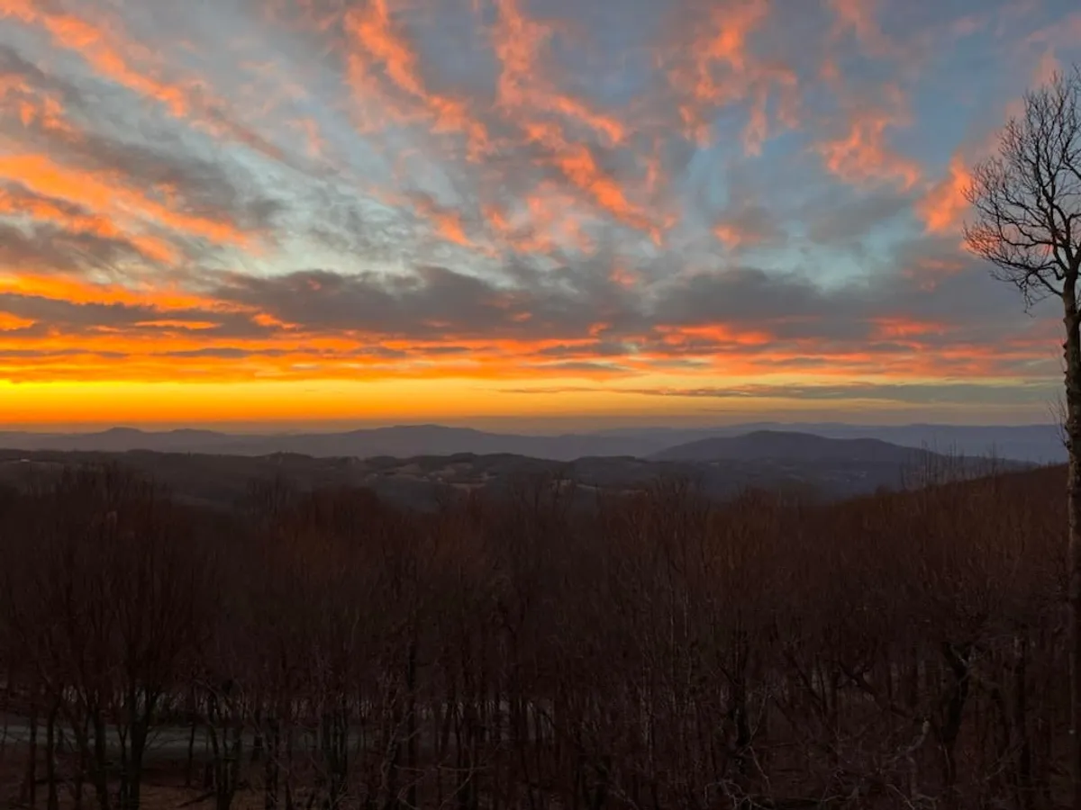A stunning view of a vibrant sunset casting warm hues over layered mountain ranges and bare trees. The mood is serene and meditative, evoking the magic of twilight moments and the grandeur of the natural setting.