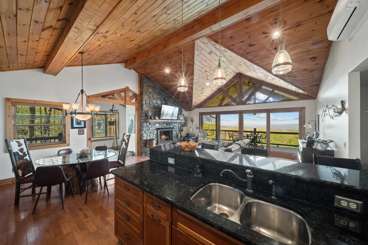 The image presents a warmly lit open-concept living and dining area with a kitchen island, dining table, a stone fireplace, and large windows offering a scenic view. The space is furnished with a blend of rustic and modern elements, showcasing the heart of the home's communal area.