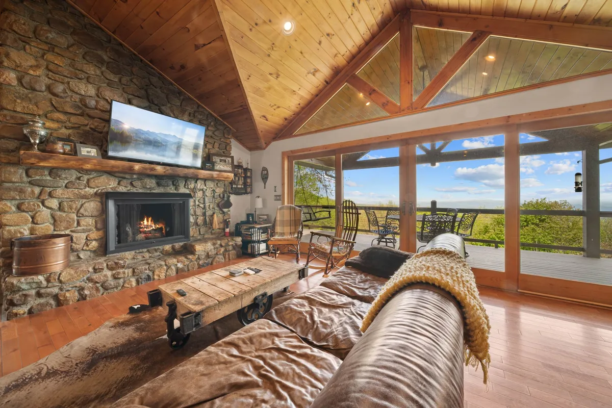 A cozy living area anchored by a stone fireplace, surrounded by large windows that let in abundant natural light and offer glimpses of mountain scenery. Rustic wooden furniture and ample seating make it an inviting spot for gatherings.