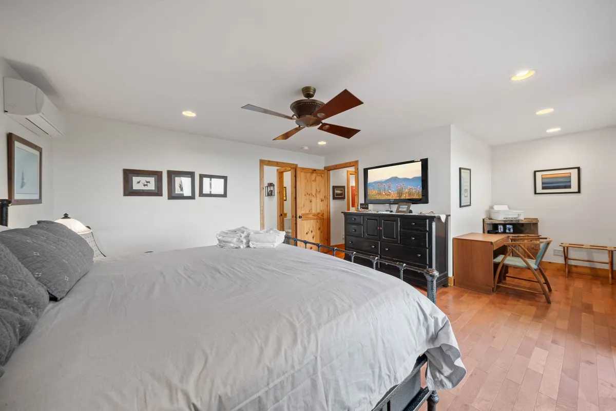A serene king-sized bedroom with a plush gray bed, wooden dresser, television, and a wooden desk and chair. Framed artwork adorns the walls, and soft, natural light fills the space, emphasizing its comfort and tranquility.