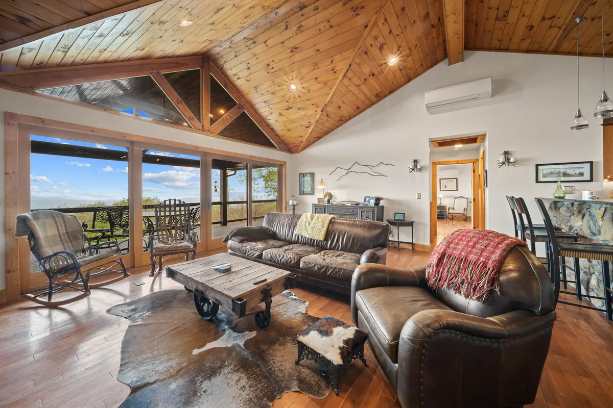 A bright and spacious living area with comfortable leather sofas, a rustic coffee table, large windows framing scenic views, and tasteful decorative wall art. The setting encourages relaxation, conversation, and togetherness.