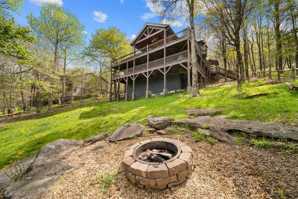The image showcases an inviting two-story mountain home nestled among lush trees, with a cozy fire pit surrounded by chairs in the foreground. The setting highlights the home's connection to nature and the opportunities for guests to relax outdoors, whether gathered around the fire pit or soaking in the expansive mountain views from the deck.