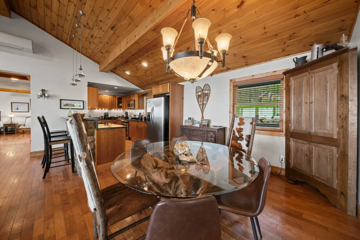 A welcoming view into a kitchen and dining area with a wooden dining table under a stylish chandelier, a rustic wooden ceiling, and an adjacent kitchen area. The layout encourages gatherings for meals and memorable celebrations.