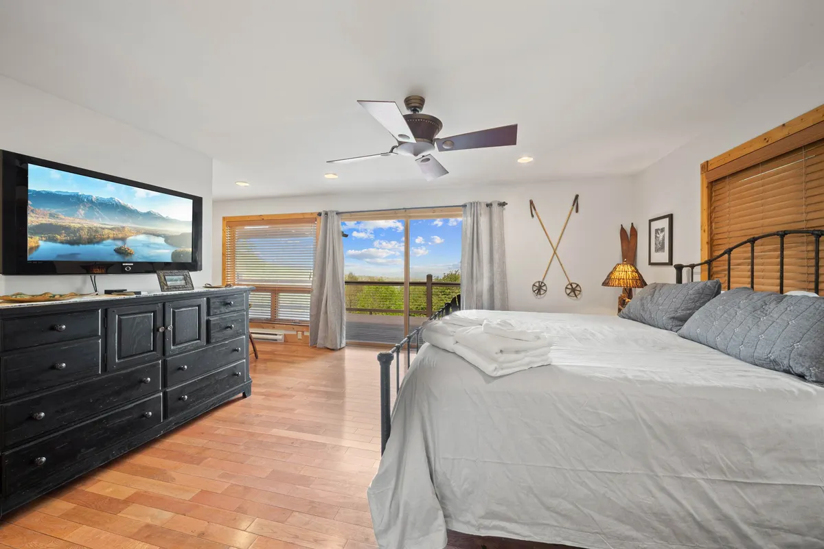 A peaceful queen-sized bedroom with gray linens, a large window offering a scenic view, a black dresser with a TV, and tasteful wall art. The room feels calm and relaxing, perfect for unwinding after a day outdoors.
